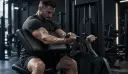 Mand siddende i en preacher curl-maskine med overarmene på en skrå pude, hænderne om maskinens håndtag, vægtmagasin synlig bagved